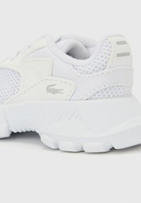 Biela športová obuv s hornou časťou zo sieťoviny, textúrovanými detailmi a gumovou podrážkou. Dizajn obsahuje jemné prvky značky a aerodynamický tvar.