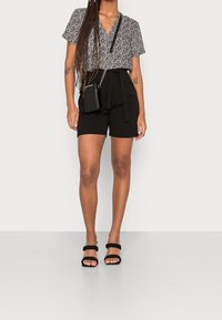 Donna che indossa pantaloni corti neri a vita alta con una cintura, una blusa a maniche corte con stampa, sandali neri e una borsa a tracolla nera.