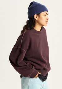 Felpa oversize color marrone con polsini e colletto a coste, abbinata a un berretto di maglia blu navy. Modello che indossa jeans azzurri.