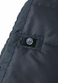 Ciemnoszary materiał z wypełnieniem o gładkiej teksturze, z czarnym paskiem i metalowym detalem w postaci guzika. Widoczne skośne szwy na krawędzi.