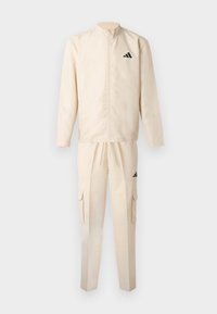 Μπεζ φόρμα που περιλαμβάνει ζακέτα με φερμουάρ και παντελόνι με ελαστική μέση, και τα δύο με μαύρες λωρίδες ως διακοσμητικά στοιχεία και τσέπες τύπου cargo στο παντελόνι.