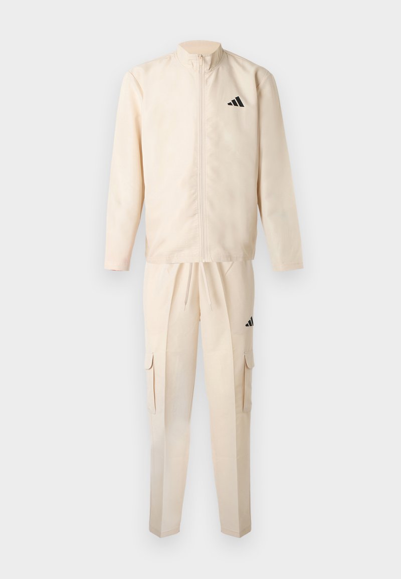 Survêtement beige comprenant une veste zippée et un pantalon à taille élastique, tous deux ornés de bandes accent noires et de poches cargo sur le pantalon.