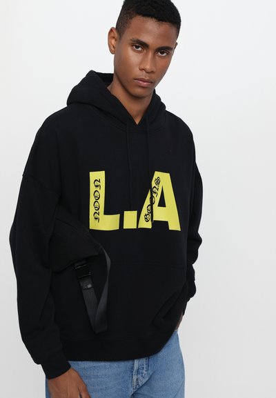 Hoodie noir avec de grands designs en jaune "L.A." et du texte. Présente une poche kangourou et une capuche avec cordon. Porté avec un jean bleu clair.