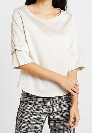 Lichtbeige blouse met een ronde halslijn en driekwart trompetmouwen met knoopaccenten. Draagt met zwart-witte geruite broeken.