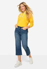 Sudadera amarilla con cordones blancos, combinada con jeans recortados azules y zapatillas blancas. Presenta un corte relajado y un look casual.