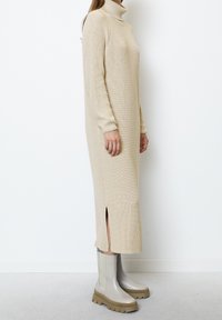 Femme portant une robe midi en tricot beige à col roulé avec des fentes latérales et des bottines gris clair à semelles épaisses, debout devant un fond blanc.