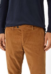 Bruine corduroy broek heeft een geribbeld textuur, voorzakken en een zwarte knoopsluiting. Gepaard met een marineblauw shirt met lange mouwen.