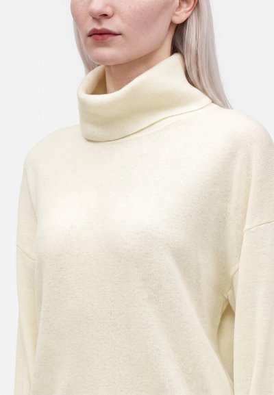 Gros plan d'une personne portant un pull à col roulé crème à manches longues et des cheveux blonds clairs et raides.