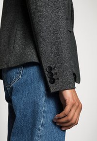 Grau gemusterter Blazer in Fischgrätoptik mit drei schwarzen Knöpfen am Ärmel, kombiniert mit blauen Denim-Jeans, die einen strukturierten Entwurf und eine strukturierte Textur zeigen.