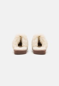 Chaussons en fausse fourrure beige avec une texture douce, ouvert à l'arrière et doté d'une semelle en caoutchouc marron. Forme arrondie et design minimaliste.