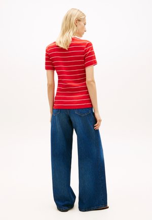 Vaaleahiuksinen nainen, jolla on punavalkoraidallinen lyhythihainen paita ja leveälahkeiset siniset farkut, seisoo valkoista taustaa vasten selin.