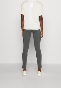 Ljusbeige t-shirt med tre vita ränder på baksidan, i kombination med grå leggings med svarta sidostriped och krämfärgade träningsskor.