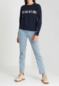 Navy long-sleeve shirt with white text, light blue jeans, and beige sneakers with a textured sole. Casual outfit with modern design.