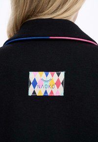 Blond vlasy na čiernom odeve s modrými a ružovými detailmi, s farebným obdĺžnikovým nášivkom s diamantovým vzorom a prešitým textom "NAOKO" a dizajnom rias.