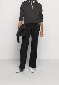 Person in einem dunklen Kapuzenpulli mit gesticktem Logo, schwarzen Hosen und weißen Turnschuhen, der eine gefaltete schwarze Steppjacke vor einem schlichten Hintergrund hält.