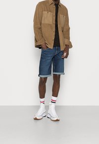 Camisa de pana color marrón con dos bolsillos frontales, camiseta negra, pantalones cortos de mezclilla azules y zapatillas altas blancas con suelas marrones y calcetines a rayas.