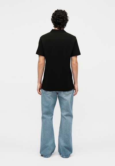 Personne aux cheveux bouclés vue de dos, portant un polo noir à manches courtes et un jean bleu clair ample.