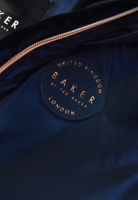 Ciemnoniebieska tkanina z złotym zamkiem błyskawicznym i okrągłą czarną naszywką z napisem "United Kingdom Baker by Ted Baker London."