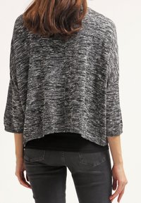 Mujer con cabello castaño ondulado de longitud media lleva un suéter gris suelto y texturizado sobre una camiseta negra y jeans gris oscuro, vista desde la espalda.