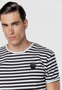 Koszulka w czarno-białe paski z krótkimi rękawami, okrągłym dekoltem oraz czarną okrągłą naszywką z detalem nuty muzycznej.