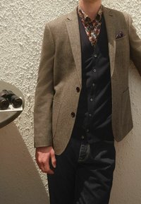 Brun høringsbåndsblazer med mørke knapper over en blomsterbluse og en navy vest, parret med mørke jeans. Kikkerter på et rundt bord i nærheden.