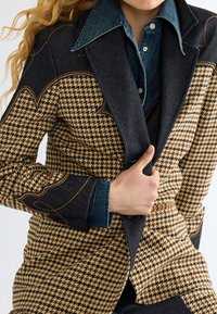Άτομο που φοράει σακάκι με σχέδιο houndstooth και σκούρα τζιν πέτα και γιακά πάνω από σκούρο μπλε πουκάμισο με κουμπιά, κρατώντας το σακάκι κλειστό.