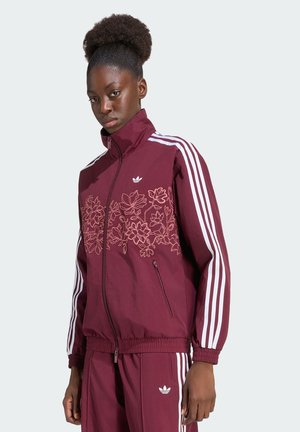 Kobieta o ciemnej karnacji i kręconych włosach, ubrana w bordowy dres Adidas z białymi paskami i kwiatowym haftem na kurtce.