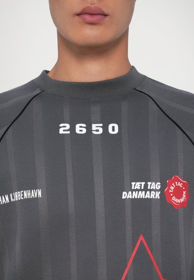 Close-up de uma pessoa a usar uma camisa listrada cinza-escura com o texto branco "2650," "TÆT TAG DANMARK," e um logótipo circular vermelho no peito.