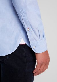 Helles blaues Hemd mit strukturiertem Stoff, kontrastierenden roten Nähten an den Manschetten und einem abgerundeten Saum. Kombiniert mit dunkler Jeans.