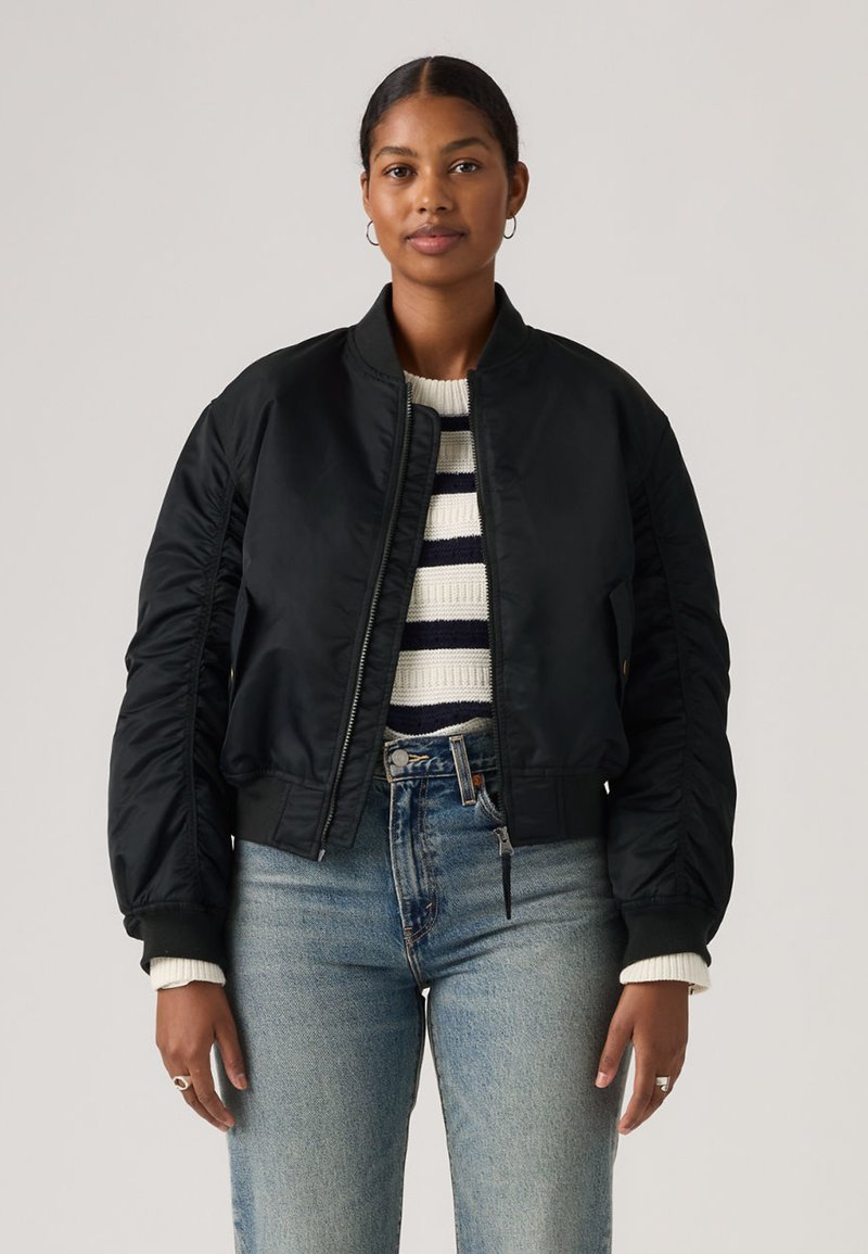 Donna che indossa una giacca bomber nera sopra un maglione a righe bianche e blu navy, abbinati a jeans azzurri, su uno sfondo semplice.
