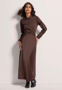 Bruine jurk met lange mouwen, een kraag, knopen aan de voorkant en een aansluitende taille, gecombineerd met zwarte enkellaarsjes. Soepele structuur.
