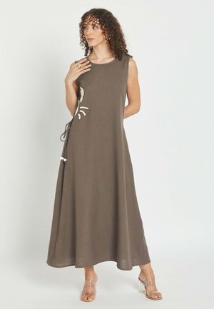 Mujer con cabello rizado que lleva un vestido midi marrón sin mangas con detalle de lazo lateral y sandalias de tacón alto con tira transparente, de pie.