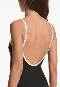 Fekete bodysuit alacsony hátrészszel, vékony krémszínű pántokkal és sima textúrával. Figyelemre méltó varrási részletek a pántok csatlakozásánál.