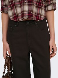 Personne portant une chemise écossaise rouge et beige raccourcie, rentrée au-dessus d'un pantalon noir taille haute, tenant un sac à main tissé marron dans la main gauche.