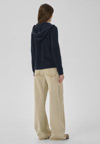 Marineblauer Kapuzenpullover aus weichem Stoff kombiniert mit hellbeigen, weitem Hosen mit sichtbaren Nähten und lässiger Passform. Rückansicht des Outfits.