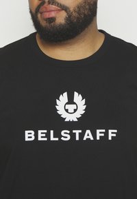 Juodas medvilninis marškinėlis su apvaliu kaklu ir baltu "BELSTAFF" logotipu bei viršuje esamu grafiniu emblemu, derinamu su medžiagos tekstūra.