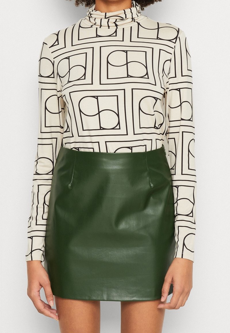 Maglietta a maniche lunghe beige con motivi geometrici neri, abbinata a una mini gonna in pelle verde aderente con tasche laterali.