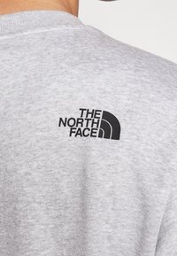 Šedá bavlněná mikina s černým logem The North Face na horní části zad. Materiál má hladkou texturu a design s kulatým výstřihem.