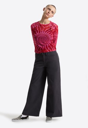 Model mit kurzen Haaren trägt ein rotes Langarmshirt mit pinken Spiralmustern, weite schwarze Hosen und schwarz-weiß karierte Slip-On-Schuhe.