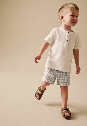 Lächelndes Kleinkind geht, trägt ein weißes kurzärmeliges Hemd mit Knöpfen, graue karierte Shorts und braune Sandalen vor beigefarbenem Hintergrund.