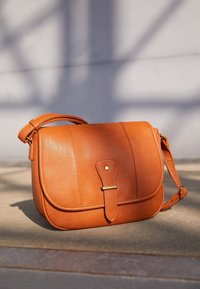 Tan læder skuldertaske med en buet klap, messingbeslag og to dekorative sømme på fronten. Justerbar reim inkluderet.