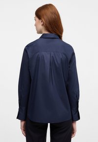 Mørkeblå skjorte med krave, lange ærmer og plisseret detalje bagpå. Glat stof med knaplukning ved ærmerne.