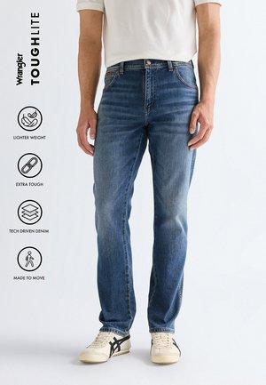 Uomo con jeans in denim blu e sneaker bianche in piedi su sfondo bianco, con icone dei benefici del prodotto Wrangler Toughlite a sinistra.