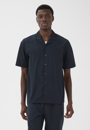 Homme portant une chemise à manches courtes bleu marine foncé à boutons avec col et un pantalon assorti, debout devant un fond clair uni.