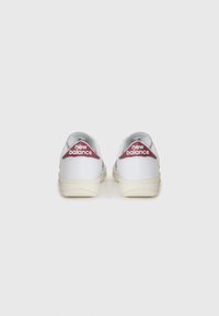 Hvite lær sneakers med lav topp-design, som har et rødt stoffmerke på hælen med "new balance"-logo, gummisåle med strukturert grep.