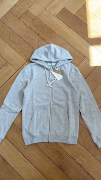 Hoody cinza claro em tecido suave, com capuz ajustável por cordão, duas bolsos frontais e uma etiqueta presa na gola.