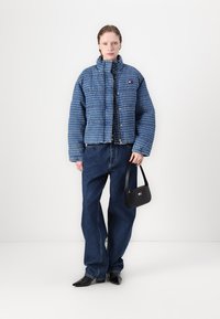 Blauwe pufferjas met een textuurafwerking, gecombineerd met wijde blauwe jeans. Houdt een kleine zwarte handtas vast; puntige zwarte schoenen maken de look compleet.