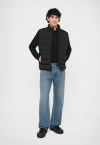 Chaleco acolchado negro con costuras horizontales, cierre de cremallera y cuello alto, superpuesto sobre una camiseta negra y combinado con jeans de pierna ancha azules y zapatillas negras.