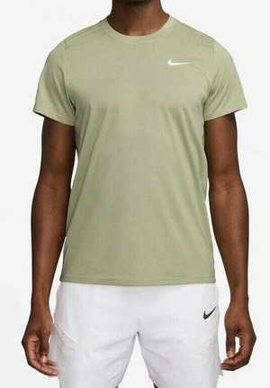 Mann trägt ein hellolivgrünes kurzärmliges Nike-Shirt und weiße Nike-Shorts, steht mit entspannten Armen an den Seiten vor einem einfarbigen Hintergrund.