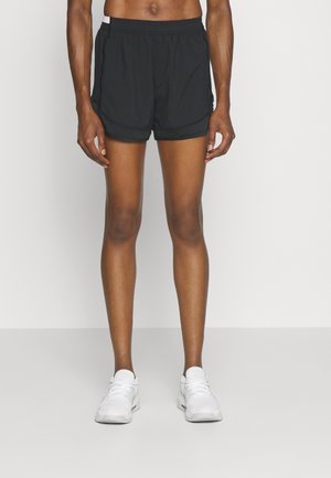 Athletische Person, die schwarze Laufsportshorts und weiße Turnschuhe trägt, steht vor einem schlichten hellen Hintergrund.